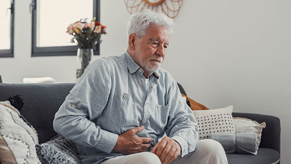 Elderly man sits on a sofa holding his stomach in pain. He strug