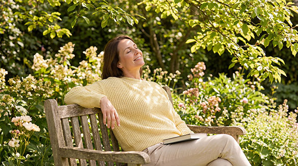 Middle aged woman relaxing on a wooden bench in a sunny garden. Happy female sitting outdoors with a book on her lap. Wellness and mindfulness concept